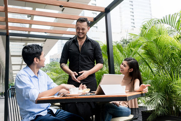 Friendly young waiter talking with the clients after serving them coffee and food at a table...