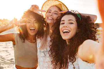 Three joyful multiethnic women 20s in summer clothing smiling at camera, while taking selfie photo...