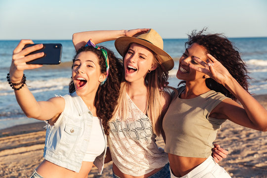 Three joyful beautiful women 20s with different color of skin wearing summer clothing smiling at camera, while taking selfie photo on smartphone during walk at seaside