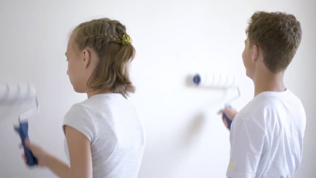 Teen boy and girl makes repairs in new flat. Sister and brother painting wall in apartment room. Children paints the wall, using a rollers. Young friends laughing and having fun. 