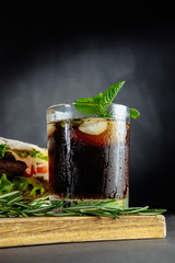 Glass of cola with ice, mint and  rosemary on black background  big sandwich, street food
