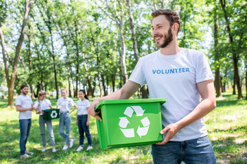 young male volunteer holding recycling box in park with friends on background