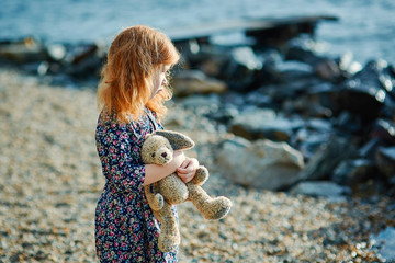 Lonely sad child stands on the shore, hugs the toy and looks into the distance