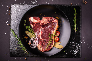 Raw meat, a beef steak, pork or veal, lamb, lies on a black ceramic plate, on a black slate. Next to the meat are spices and vegetables. Top view. Copy space