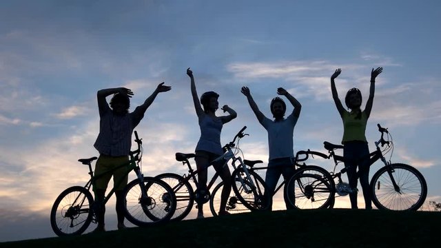 Four cheerful cyclists dancing at sunset sky. Four yung friends with bikes having fun on evening nature background.