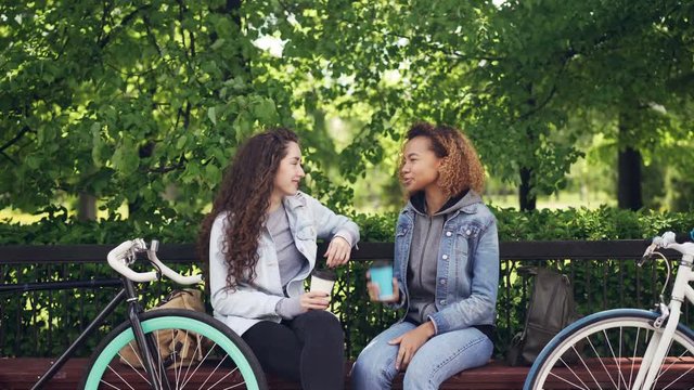 Attractive young women Caucasian and African American are chatting and drinking takeaway coffee while sitting on bench in park and resting after riding bikes.