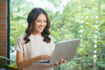 Portrait of young woman working holding laptop standing against panoramic window with garden view