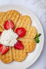  Norwegian heart shaped waffles topped with strawberries,  mint and whipped cream on white plate and white wooden background. St. Valentine's Day breakfast. Romantic breakfast. Copy space