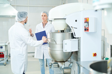 Two confectionery factory workers standing in white coats and discussing while operating dough...