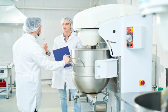 Two confectionery factory workers standing in white coats and discussing while operating dough mixing machine.