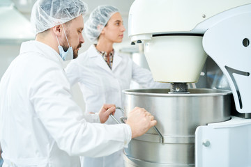 Two confectionery factory employees in white coats working with dough mixing machine.