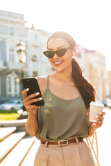 Photo of smiling brunette woman wearing casual summer outfit and wireless earpod, standing in sunlit street with takeaway coffee and black mobile phone