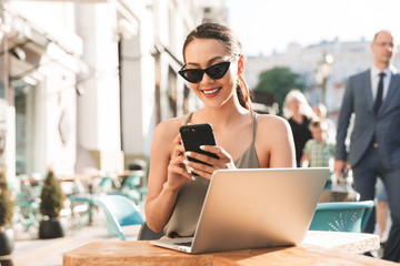 Photo of caucasian businesswoman wearing black sunglasses and dress holding cell phone, while sitting in cozy cafe or restaurant outside in summer