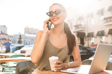 Photo of caucasian businesswoman wearing black sunglasses and dress talking on mobile phone, while resting in street cafe or restaurant in summer