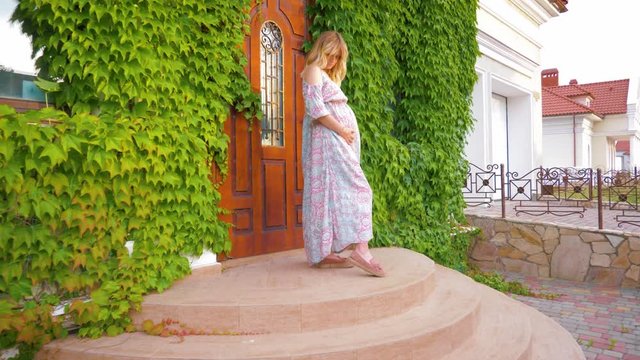A happy pregnant girl is standing on the steps of a new rich house against the backdrop of green thickets and stroking her belly in anticipation of a miracle.