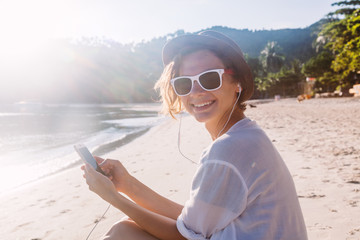 Young beautiful woman hipster traveler in headphones and with mobile phone in hands, on the beach in the sun