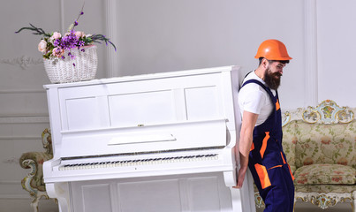 Loader moves piano instrument. Man with beard, worker in overalls and helmet lifts up piano, white...
