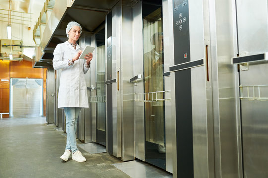 Confectionery factory employee in white coat standing near elevators and using tablet computer. 
