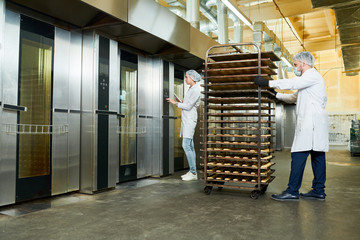 Confectionery factory employees in white coats waiting for elevator to transport trays rack with...