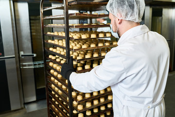 Back view of confectionery factory employee in white coat transporting trays rack with pastry. 