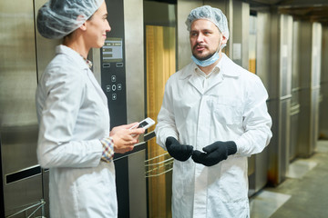 Confectionery factory workers in white coats standing near elevators and talking. 
