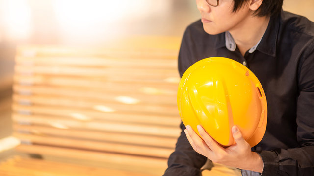 Young Asian engineer man holding personal yellow protective safety helmet while sitting on wooden bench. Male architect, Construction and Engineering concepts