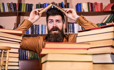 Deadline concept. Man on shocked face between piles of books, while studying in library, bookshelves on background. Teacher or student with beard sits at table making roof out of book, defocused.