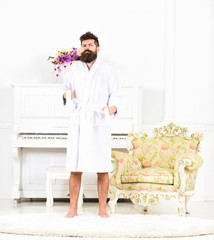 Full length portrait of a young man in a bathrobe standing isolated in white room with piano. Bearded guy tightening his robe. Sleepy hipster in his apartment