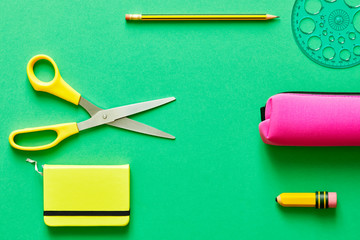 School tools such as yellow scissors, pencil, ruler, eraser, and pencil case, over a green flatlay...