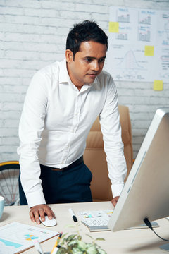 Mid adult Indian businessman standing at office desk and working on computer