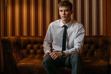 A young, handsome man in a business suit is sitting on the brown leather sofa