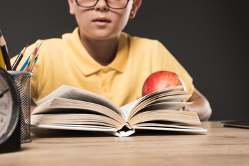 cropped image of schoolboy in eyeglasses eating apple and reading book at table with colour pencils and clock on grey background