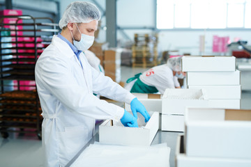 Side view of confectionery factory worker standing in white coat and preparing empty paper box. 