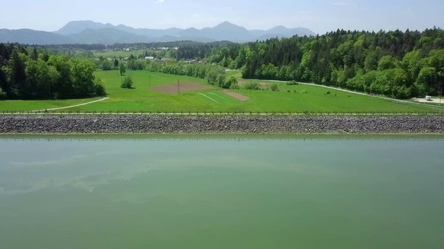 Aerial: Flying over green lake towards dam on it on sumer sunny day. Flying with drone and filming on beautiful sunny summer day.