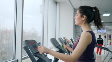 healthy lifestyle, sportswoman builds up muscles using Elliptical trainers in fitness club with large panoramic windows