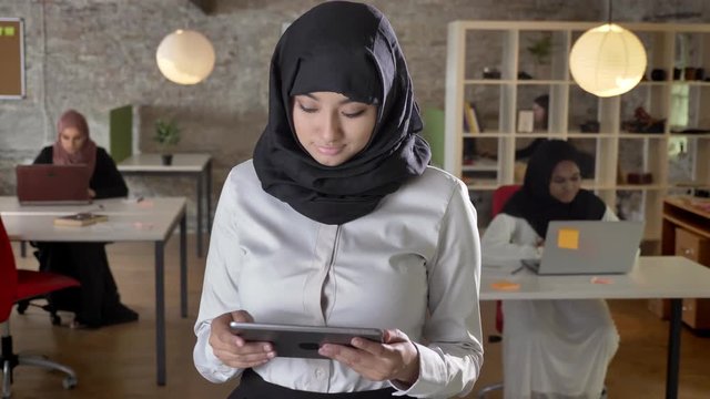 Young muslim women in hijab using tablet, looking in camera, smiling, business womens typing on laptop in modern office