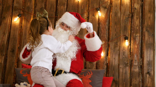 Cute little girl pulling Santa's beard to check if it's real sitting on his lap