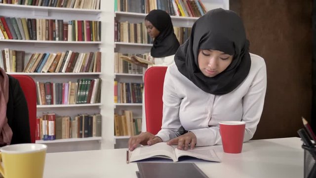Three young muslim womens in hijab reading books in library, studying for exam, shelves with books background, islamic students