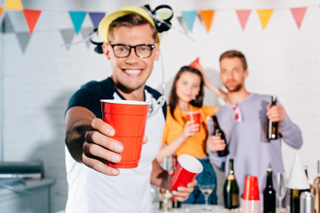 close-up view of smiling young man holding plastic cup with beer while friends partying behind