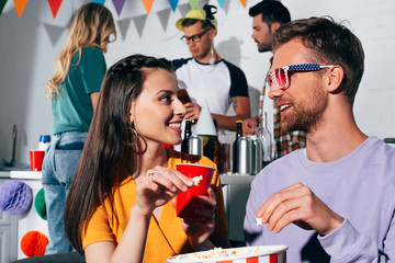 happy young man and woman smiling each other while drinking beer and eating popcorn at home party