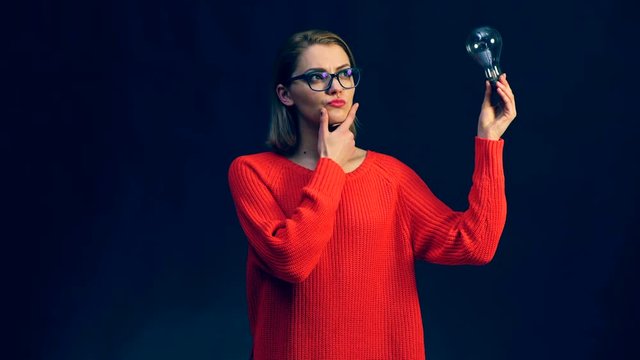 Thinking woman in glasses looking up with light idea bulb in hand isolated on black background. Idea. Discovery. PHD.