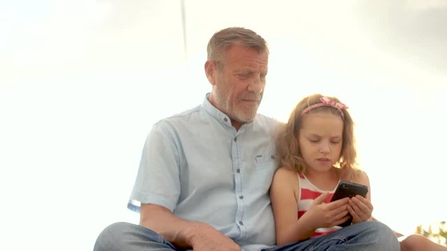 Mature man and little girl curly schoolgirl watching video in smartphone. Against the blue sky. Father's Day. Family day