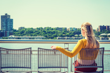 Fototapeta na wymiar Empty Chairs for You. lonely girl waiting friendship. Wearing yellow long sleeve fit crop T shirt, young woman with long blonde hair sitting on chair by Hudson River in New York, facing New Jersey.