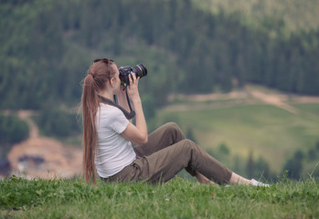 Girl with camera sits on a hill and photography nature. Summer day