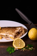 Cutted lemonwith black knife and plate with Grilled fish in the sauce and with spices and herbs on a wooden background