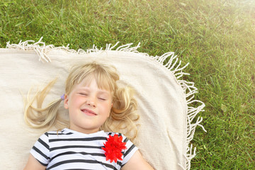 Little cute blond girl lying on blanket over green grass lawn and smiling. Adorable child having fun outdoors. Happy family, parenthood and hildhood concept