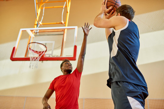 African-American man trying to stop contender from throwing ball into basket while playing basketball in gym. 