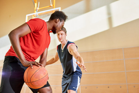 Young sportsman trying to interrupt black man dribbling ball while playing basketball in gym. 