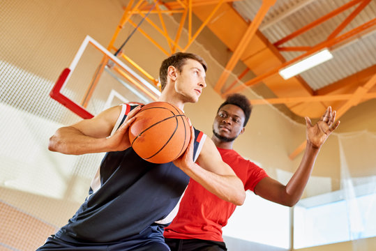 Young black man trying to stop contender from shooting goal while playing basketball together in gym.