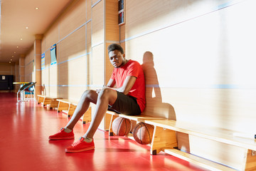 Handsome African-American man in basketball uniform looking at camera while sitting on bench in gym.